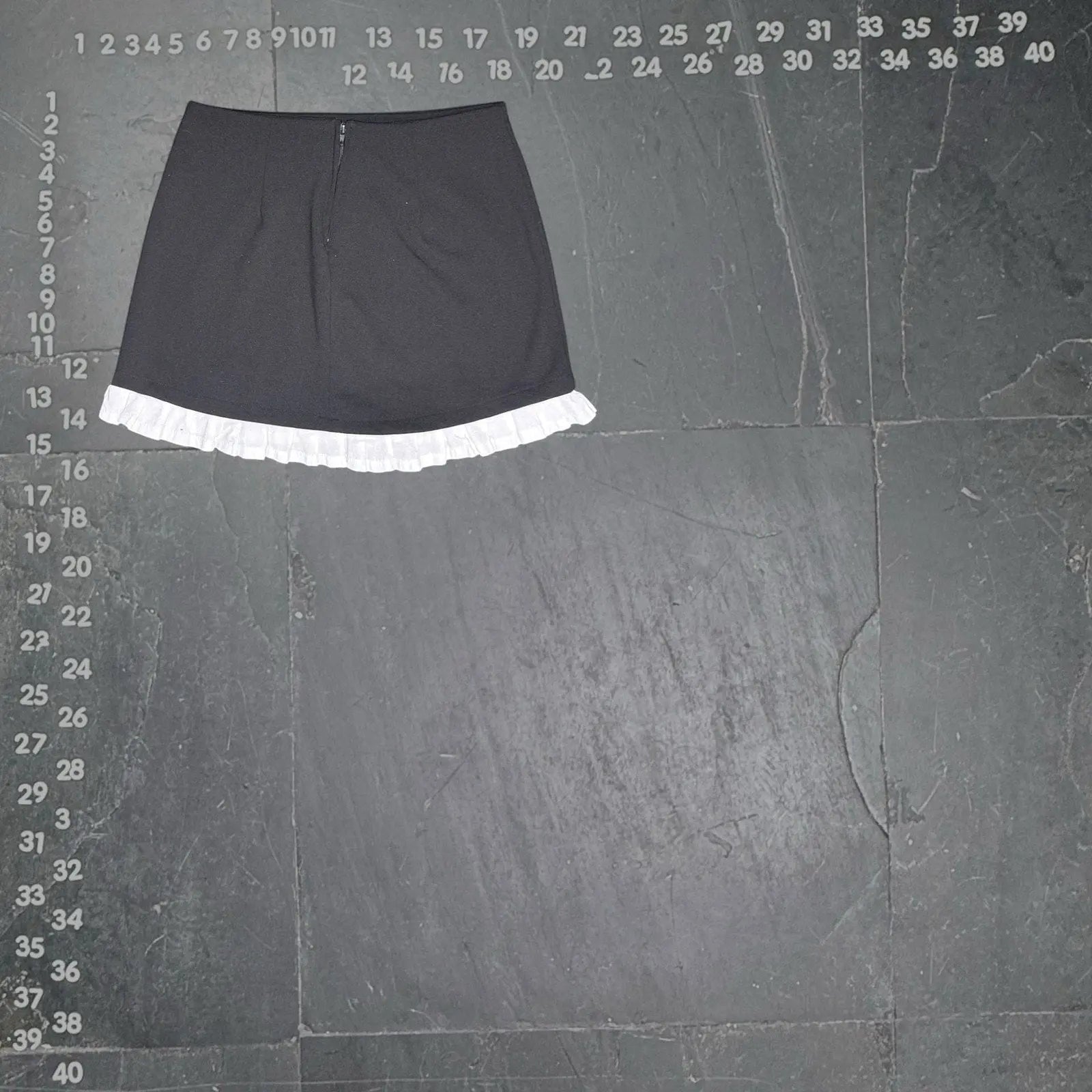 The Room Collective black mini skirt with white ruffled hem. High-waisted, slim fit, polyester/spandex blend with cotton ruffle. Back zipper.