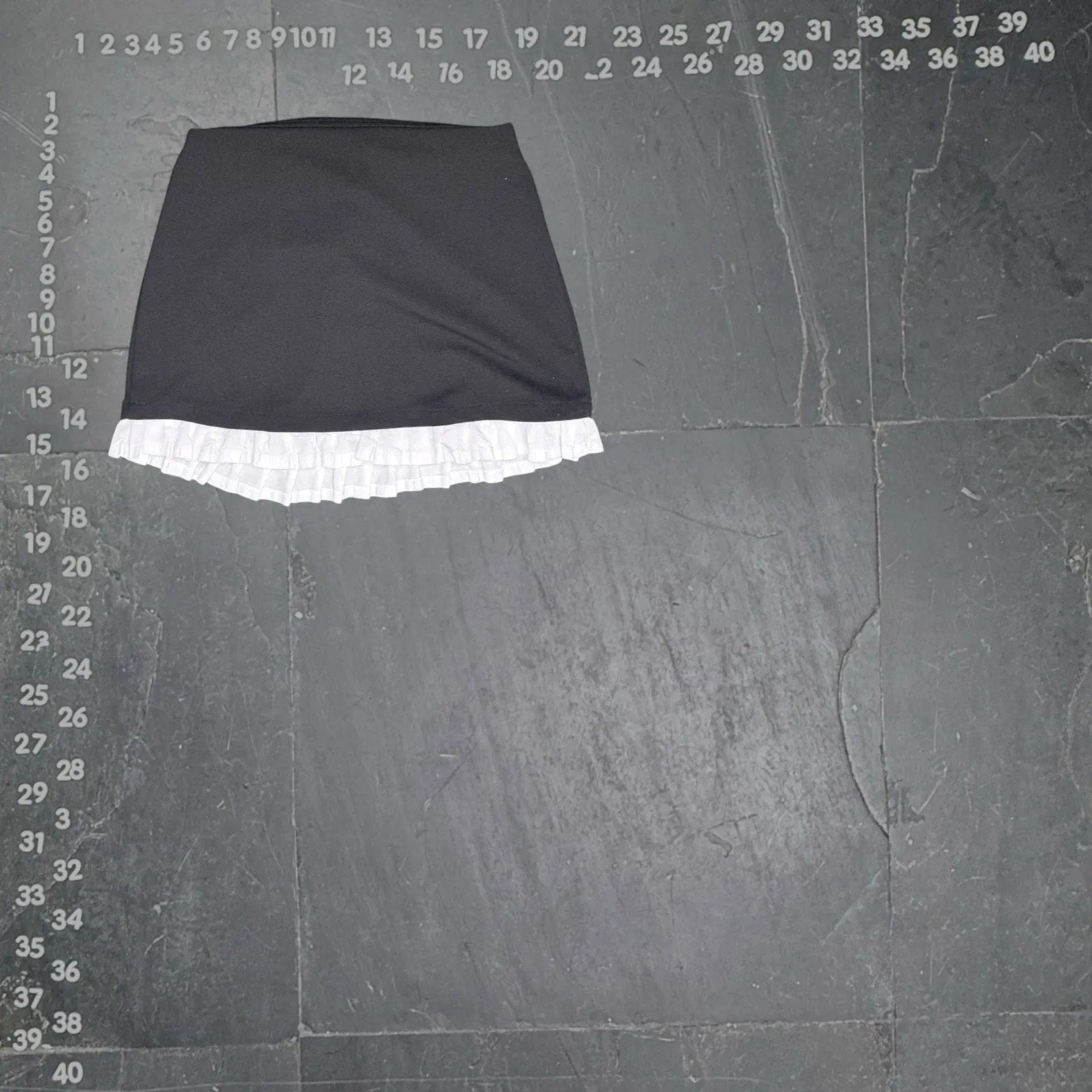 The Room Collective black mini skirt with white ruffled hem. High-waisted, slim fit, polyester/spandex blend with cotton ruffle. Back zipper.